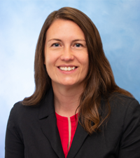 A smiling woman with long brown hair wears a black blazer over a red top against a soft blue background.